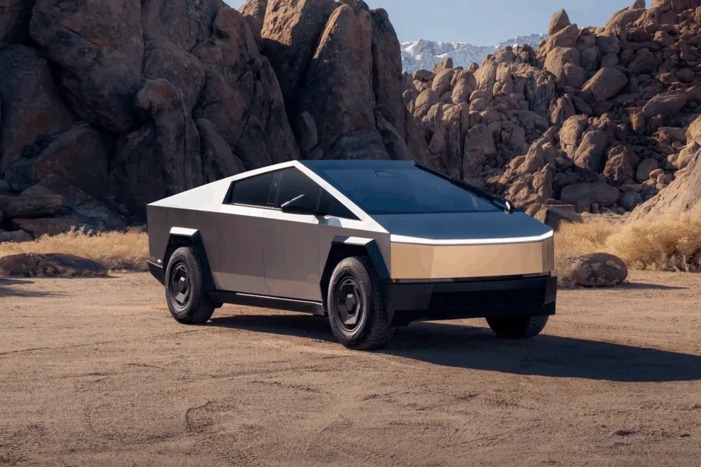 Stainless steel Tesla Cybertruck parked on desert dirt among rocky boulders and distant snowy peaks