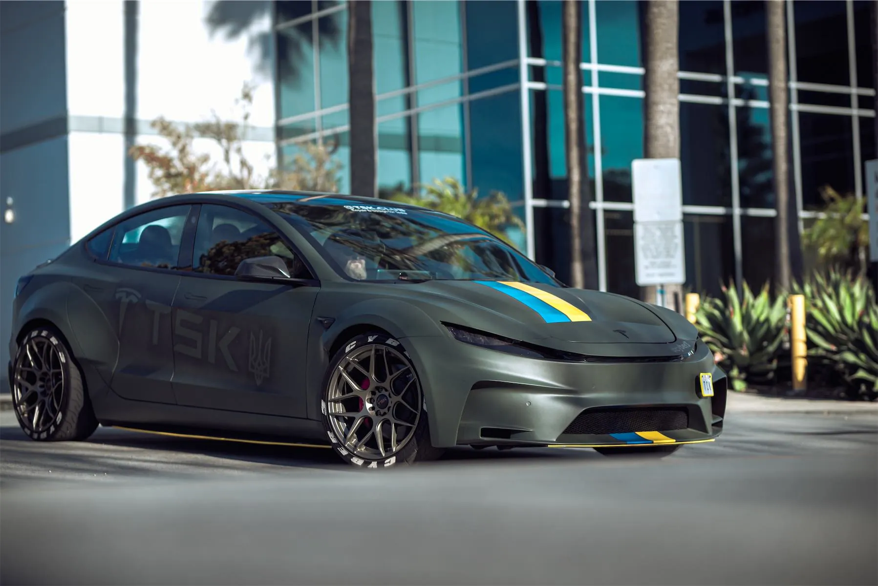 Cyber 3 project: matte green Tesla Model 3 widebody with blue and yellow stripes by a glass office building