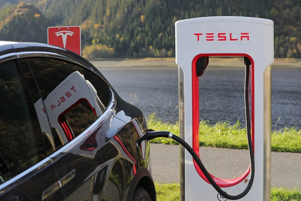 Black Tesla plugged into Supercharger station beside a lakeside road