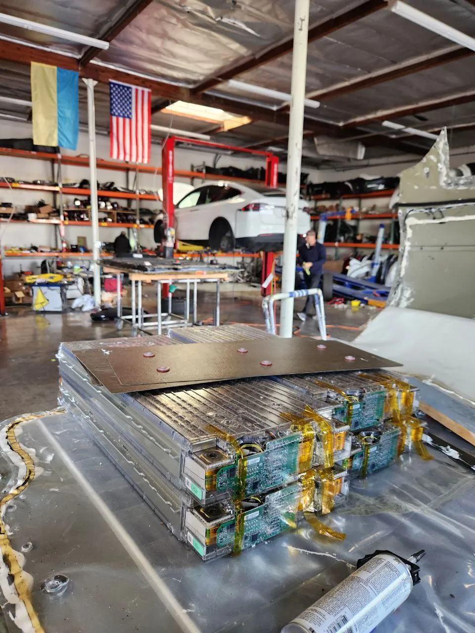 Disassembled Tesla HV battery module on shop floor with white Tesla on lift behind