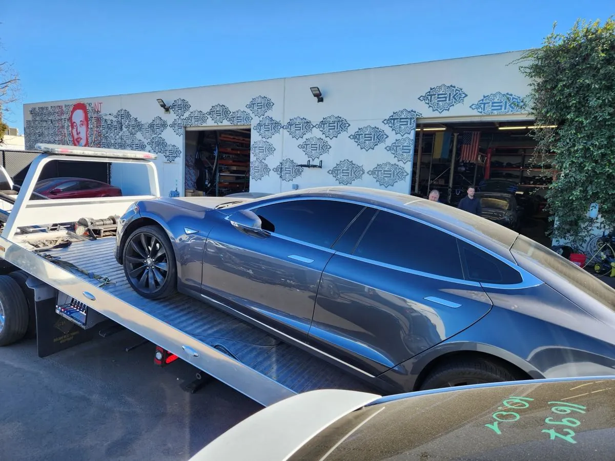 Gray Tesla Model S on flatbed tow truck outside TSK collision repair shop