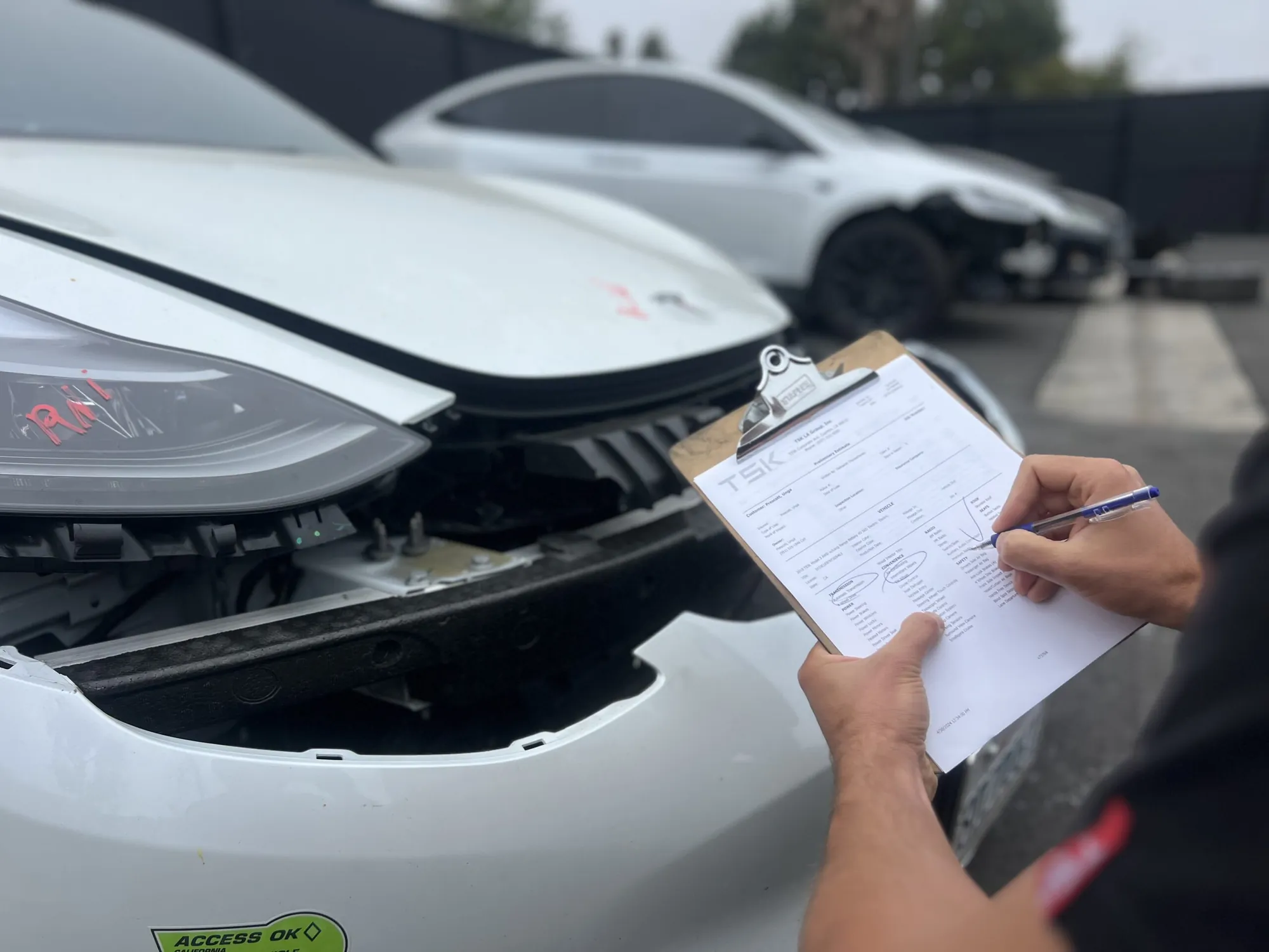 Technician documenting Tesla Model 3 front-end damage on TSK inspection form at repair shop