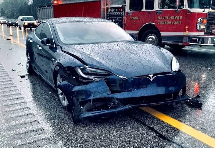 Tesla Model S with crumpled front-end collision damage on wet road near fire truck